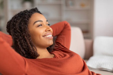 Foto de Retrato de una hermosa mujer negra sonriente inclinada hacia atrás en un cómodo sofá, feliz calma dama afroamericana descansando con las manos detrás de la cabeza, relajándose en el sofá en la sala de estar en casa, disparo de primer plano - Imagen libre de derechos
