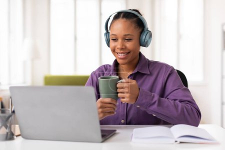 Photo for Cheerful black teenage girl in purple shirt, wearing headphones, takes relaxing break with warm cup of tea while studying with laptop at home - Royalty Free Image