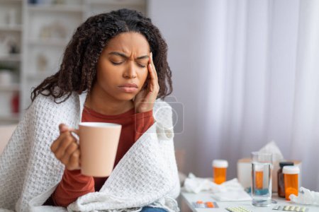 Photo for Sick young black woman suffering from headache or cold at home, african american female with pained expression sitting on couch wrapped in blanket, rubbing temples and holding cup of tea, copy space - Royalty Free Image