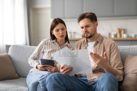 Photo for Perplexed young spouses analyzing documents together while holding digital tablet, showing concern and confusion, sitting on sofa in living room at home - Royalty Free Image
