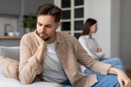 Photo for Worried man sitting on sofa with hand on cheek, deep in thought, while woman sits with her back to him, depicting relational difficulties - Royalty Free Image