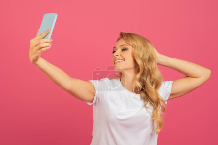Foto de Joven rubia atractiva tomando una selfie con su teléfono inteligente, posando para la foto en una camiseta blanca casual, de pie sobre el fondo del estudio rosa. Concepto de redes sociales y presencia en línea - Imagen libre de derechos