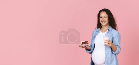 Foto de Mujer embarazada joven positiva con pastel de terciopelo rojo y vaso de leche posando aislada sobre fondo de estudio rosa, esperando que la dama sienta hambre, ansia de dulces, panorama con espacio para copiar - Imagen libre de derechos