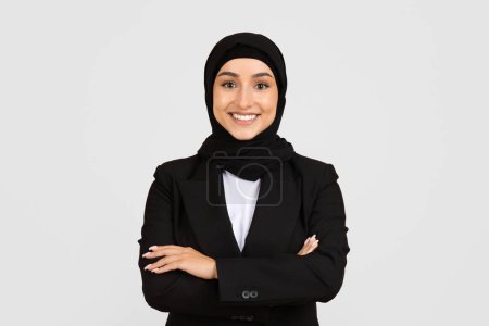 Confident and professional smiling young woman wearing hijab and black business suit with arms crossed standing on light background and smiling at camera