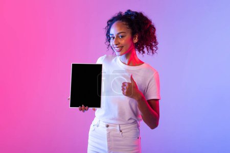 Photo for Enthusiastic black lady with curly hair smiling and giving thumbs up while holding blank tablet screen, over neon pink and purple background, perfect for digital technology marketing - Royalty Free Image