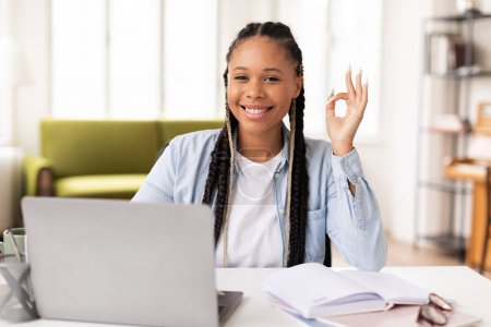 Foto de Contenido joven afroamericana estudiante americana con trenzas dando un signo de aprobación mientras estudia con el ordenador portátil en la habitación luminosa y acogedora en casa - Imagen libre de derechos