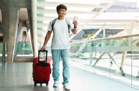 Photo for Excited young asian guy tourist with suitcases using cell phone while walking by airport, doing online checkin before boarding on flight, booking hotel or taxi or application, copy space - Royalty Free Image