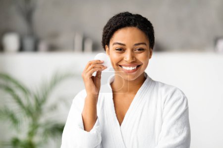 Téléchargez les photos : Joyeuse femme noire tenant un tampon de coton, prête pour sa routine de soins de la peau à la maison, jolie dame afro-américaine en peignoir blanc posant dans un cadre de salle de bain serein, regardant la caméra, copiant l'espace - en image libre de droit