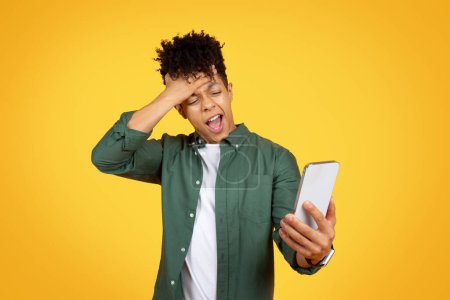 Téléchargez les photos : Stressé jeune homme afro-américain regardant l'écran du smartphone, tenant gadget et toucher le front, la lecture de mauvaises nouvelles, ont des difficultés avec l'application mobile, fond de studio jaune - en image libre de droit