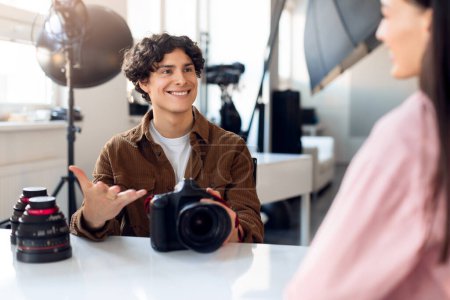 Téléchargez les photos : Jeune photographe masculin aux cheveux bouclés, tient un appareil photo reflex numérique tout en engageant une discussion légère avec un mannequin féminin, dans un environnement de studio photo ensoleillé - en image libre de droit