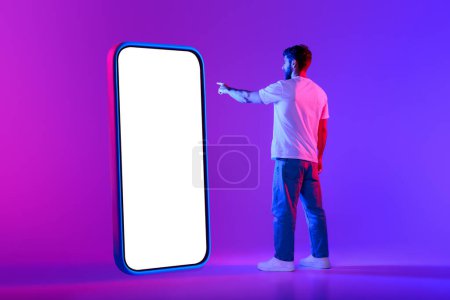 Photo for Young European man points to big cellphone screen, presenting empty mockup space for customizable content against purple blue neon lit backdrop. Online communication, applications - Royalty Free Image