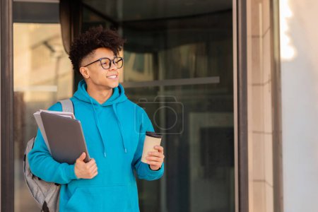 Photo for Smiling student guy in glasses casually standing at college entry door, holding takeaway coffee, laptop and workbooks outside, smiling to camera. Concept of academic pursuits and modern education - Royalty Free Image