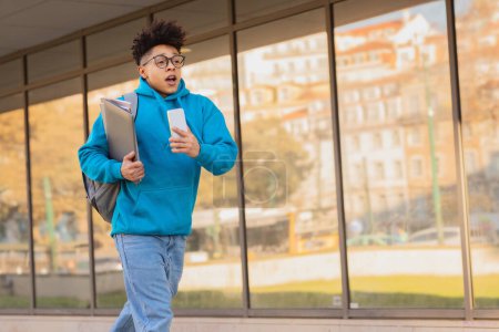 Photo for Brazilian student guy with backpack and phone rushes through the city, hurrying late for college seminars. Concept of urgency and time management challenges faced by students. Empty space - Royalty Free Image