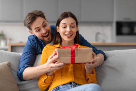 Photo for Delighted woman receiving gift with red ribbon from ecstatic man, both showing expressions of joy and surprise, comfortably seated on gray sofa in modern home interior - Royalty Free Image