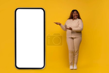 Téléchargez les photos : Positif potelée aux cheveux longs millénariste afro-américaine dame portant costume beige pointant vers un téléphone énorme avec écran blanc blanc maquette copie espace sur fond jaune, montrant l'offre en ligne - en image libre de droit