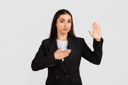 Photo for Confident young professional woman in black suit takes an oath, placing one hand on chest and other raised, standing against light gray backdrop - Royalty Free Image