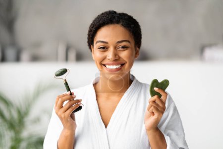 Beauty Tools. Smiling black woman holding jade guasha stone scraper and greenstone roller, beautiful african american lady looking at camera, making skincare routine, standing in bathroom at home