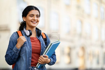 Photo for Positive young eastern lady student posing at university campus, holding exercise books, carrying backpack, looking at copy space and smiling. Education, school, college concept - Royalty Free Image