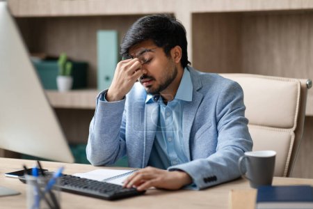 Young Hindu professional businessman wearing eyeglasses rubbing nosebridge feeling tired, experiences workplace stress while sitting at his desk with computer in modern office