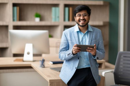 Téléchargez les photos : Homme d'affaires exécutif indien souriant avec des lunettes de navigation sur tablette numérique, se tient regardant avec confiance la caméra, tenant gadget d'ordinateur tout en posant dans le bureau moderne. Espace libre - en image libre de droit