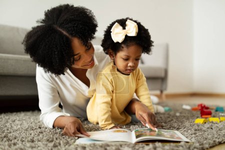 Photo for Loving young african american mother reading book with her child cute little baby girl. Mom and daughter laying on floor at home, checking colorful pictures at playbook, copy space - Royalty Free Image