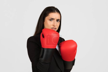 Téléchargez les photos : Inquiète femme professionnelle en colère en tenue d'affaires avec des gants de boxe rouge montrant le stress ou le défi dans un environnement compétitif, arrière-plan léger - en image libre de droit