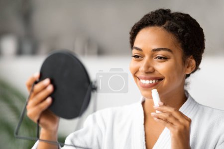 Lächelnde Schwarze Dame hält Spiegel und trägt feuchtigkeitsspendenden Lippenbalsam auf. Schöne Afroamerikanerin benutzt Chapstick und zieht hygienischen Lippenstift auf, während sie im heimischen Badezimmer steht