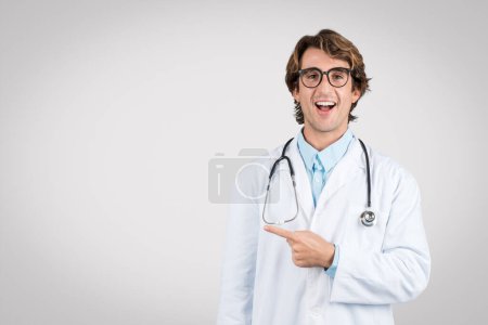 Exuberante médico varón joven en gafas y bata de laboratorio apuntando al espacio libre, encarnando profesional sanitario amigable y accesible, fondo gris