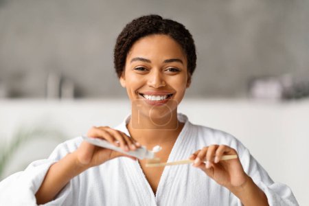 Foto de Mujer negra alegre aplicando pasta de dientes al cepillo de dientes de bambú, mujer afroamericana sonriente en albornoz preparándose para la higiene bucal en casa, de pie en el interior del baño moderno - Imagen libre de derechos