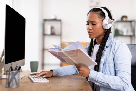 Photo for Concentrated black teen lady with braids wearing headphones, attentively reading from notebook while simultaneously working on desktop computer - Royalty Free Image