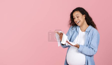 Photo for Motherhood concept. Happy pregnant woman with big belly looking at baby clothes in her hands and smiling, feeling emotional, expecting child, pink studio background, copy space - Royalty Free Image