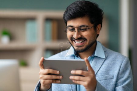 Téléchargez les photos : Homme d'affaires indien sourit tout en utilisant l'application sur tablette numérique, mélange de travail et de plaisir de la technologie dans l'environnement de bureau moderne, portrait rapproché de l'homme tenant dispositif informatique - en image libre de droit