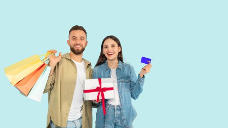 Photo for Shopping Together. Happy Young Couple With Shopper Bags Holding Gift Box And Showing Their Bank Credit Card, Advertising Great Financial Offers, On Blue Studio Background. Panorama, Free Space - Royalty Free Image