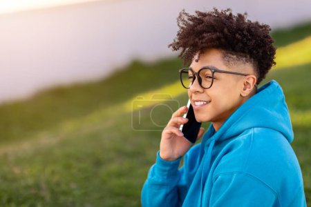 Téléchargez les photos : Vue de côté portrait d'un jeune Brésilien joyeux bavardant sur smartphone, posant dans des lunettes à l'extérieur, regardant loin avec le sourire tout en ayant une conversation téléphonique agréable. Espace vide - en image libre de droit