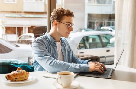 Photo for Focused caucasian young student with curly hair and glasses typing on a laptop with a pastry and coffee nearby, engaged in work at a street-view cafe table. Work, study remotely - Royalty Free Image