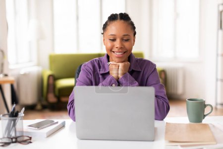 Téléchargez les photos : Contenu étudiante dame noire en chemise violette sourit comme elle a absorbé dans son ordinateur portable, étudier en ligne dans un environnement bien éclairé à la maison avec tasse de café de côté - en image libre de droit