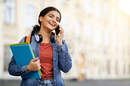 Photo for Cheerful pretty young indian woman student carrying backpack, textbooks have phone conversation with friend while walking outdoors by university campus, copy space - Royalty Free Image