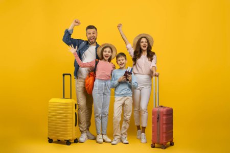 Téléchargez les photos : Heureuse famille caucasienne de quatre personnes avec valises et billets de voyage posant sur fond de studio jaune, parents voyageurs joyeux et leurs deux enfants prêts pour les vacances, profiter des voyages, copier l'espace - en image libre de droit