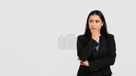 Contemplative young professional woman in black business suit looking aside at free space and touching chin, against grey backdrop, panorama, banner