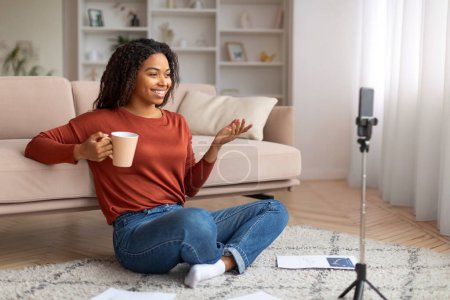 Téléchargez les photos : Heureuse jeune femme noire enregistrant un vlog sur son smartphone, s'engageant avec son public tout en tenant une tasse de café, confortablement assise sur le sol de son élégant salon, appréciant bloguer - en image libre de droit