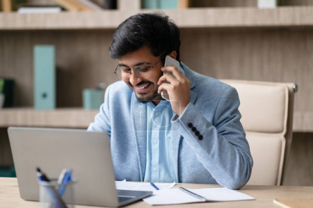 Photo for Indian professional man engages in business call, talks on cellphone at modern workplace, working online on laptop during phone conversation in office interior. Technology, career and communication - Royalty Free Image
