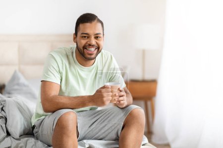 Photo for Smiling handsome bearded millennial black man wearing pajamas enjoying morning coffee, sitting on bed with cup in his hands, cozy bedroom interior, copy space - Royalty Free Image