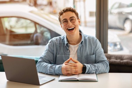 Photo for Laughing caucasian young man student with curly hair and glasses sitting with hands clasped in front of his open notebook and laptop in a bright cafe setting. Study, work - Royalty Free Image