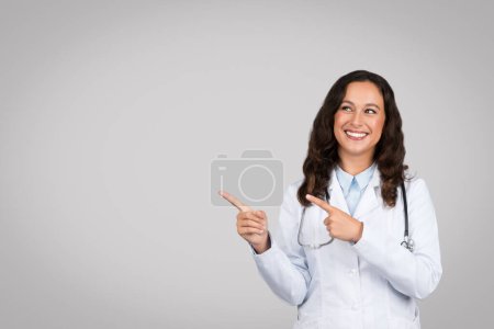 Photo for Happy young lady doctor in white coat pointing fingers up at free space, recommending medical ad and offer on grey studio background. Professional advice, health care - Royalty Free Image