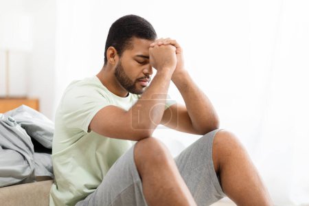 Photo for Unhappy pensive handsome african american man wearing pajamas homewear sitting on bed alone at home, suffering from depression, loneliness or anxiety, copy space. Permacrisis, midlife crisis - Royalty Free Image