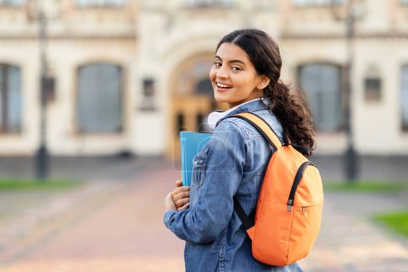Photo for Positive young woman student looking over shoulder and smiling. Cheerful eastern lady walking by city campus, carrying backpack and workbooks, copy space. Education concept - Royalty Free Image