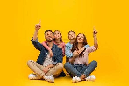 Photo for Look There. Happy Young Parents With Two Kids Sitting On Floor And Pointing Up At Copy Space, Cheerful Caucasian Family Of Four Demonstrating Free Place Above Their Heads For Advertisement Design - Royalty Free Image