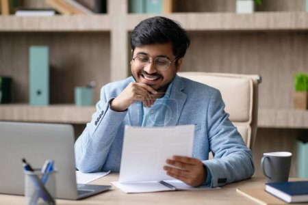 Photo for Cheerful Indian Businessman Engaging In Business Paperwork, Holding And Reading Document Near Laptop, While Sitting At Work Table In Modern Office Interior. Successful Entrepreneurship - Royalty Free Image