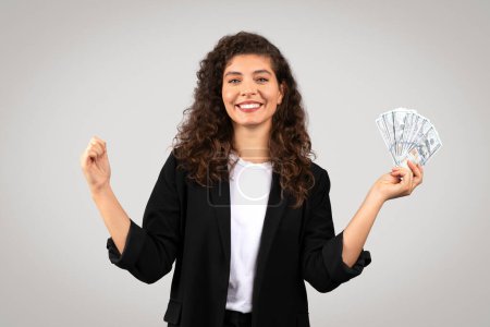 Photo for Joyful curly-haired businesswoman in black blazer holding fan of US dollar bills in one hand, celebrating financial success with fist pump and smiling - Royalty Free Image