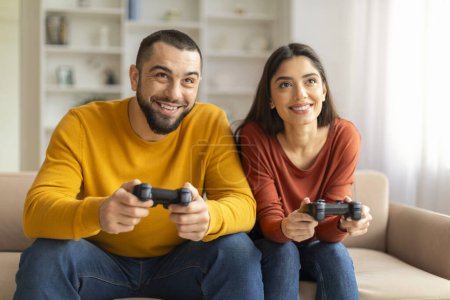 Foto de Alegre joven pareja divirtiéndose en casa, jugando videojuegos juntos, felices cónyuges millennials emocionados sosteniendo Joysticks y compitiendo entre sí mientras se sientan en el sofá en la sala de estar - Imagen libre de derechos